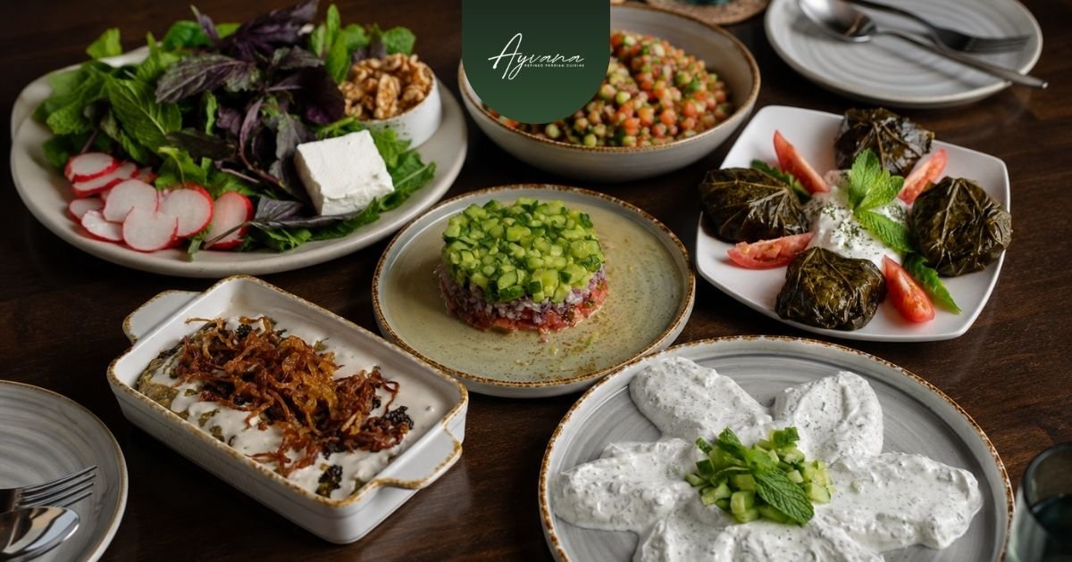 Selection of Persian appetizers for sharing, including kashk-e bademjan, mast-o-khiar, dolmeh, salad shirazi, and fresh herbs with feta arranged on a table for group dining.